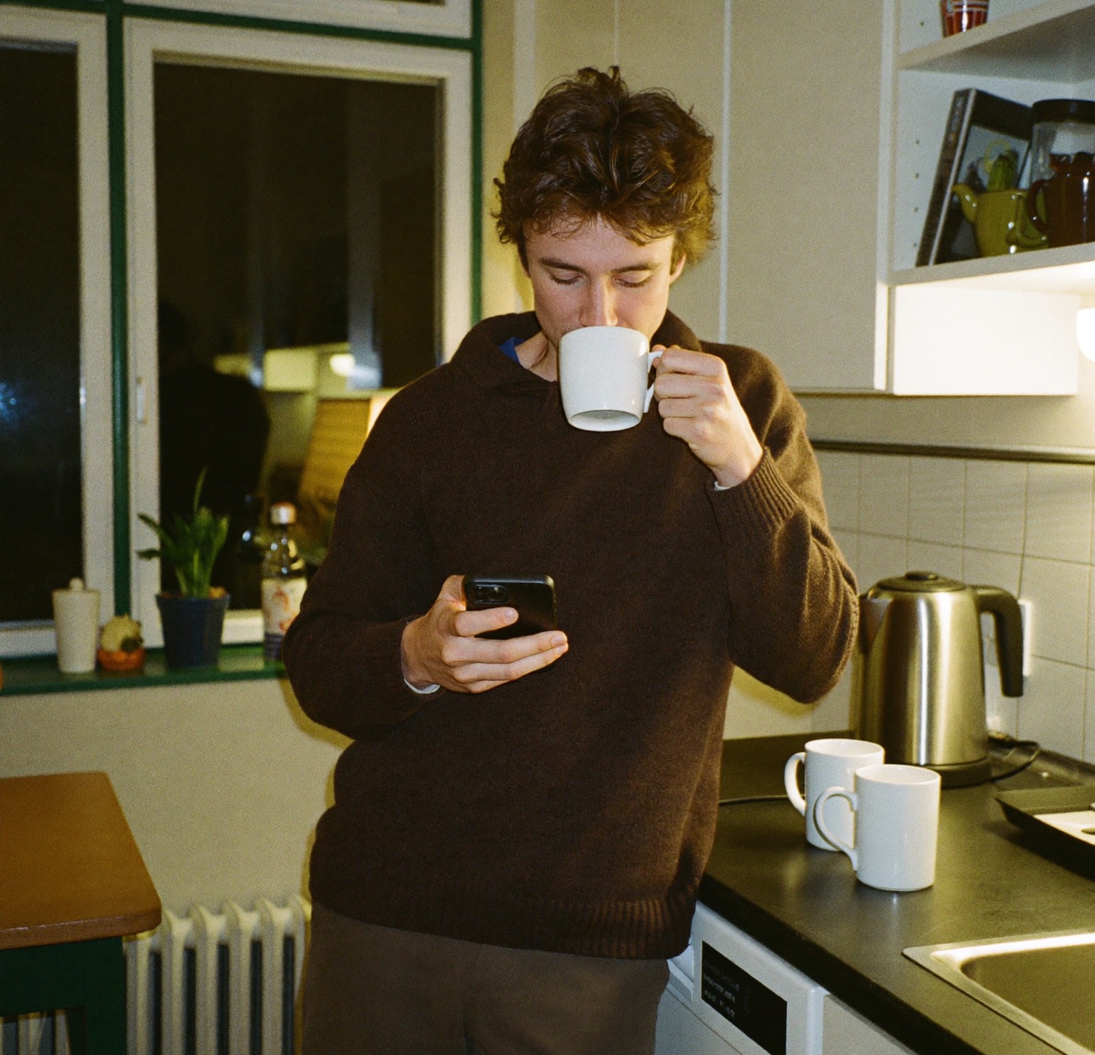 A young man drinking tea in a kitchen at night, reading his phone